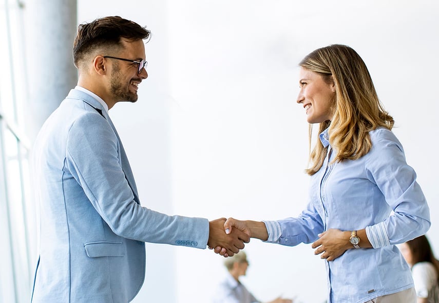 Personal Beratende der contec und con Quaesso Unternehmensberatung und Personalberatung der Gesundheitswirtschaft und Sozialwirtschaftgeben  geben sich in einem Büro die Hand, während ihr Team im Hintergrund arbeitet.