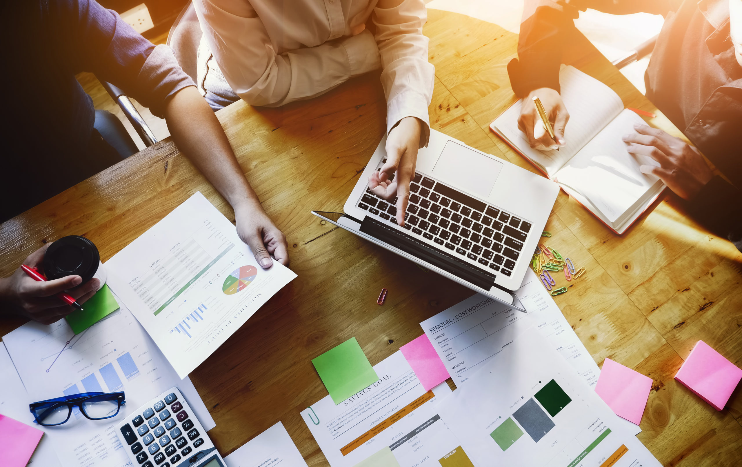 Auf einem Tisch liegen mehrere Papiere udn ein Notebook. Darauf Unterarme von Mitarbeitenden, die die Arbeitsentgelte in einer WfbM berechnen.