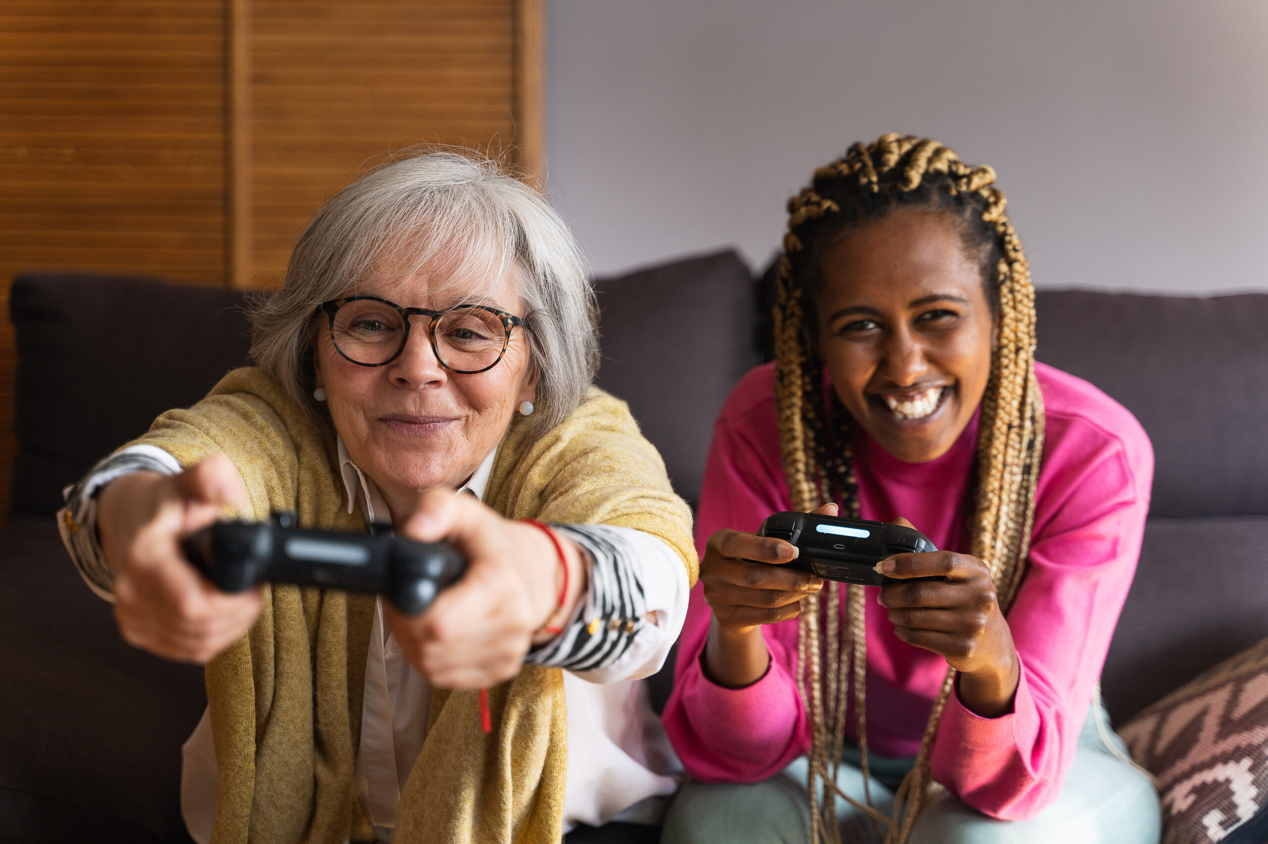 Eine junge Frau sitzt neben einer älteren Frau auf der Couch und beide halten eine Spielekonsole in der Hand. Ein Symbolbild für eine Caring Community.